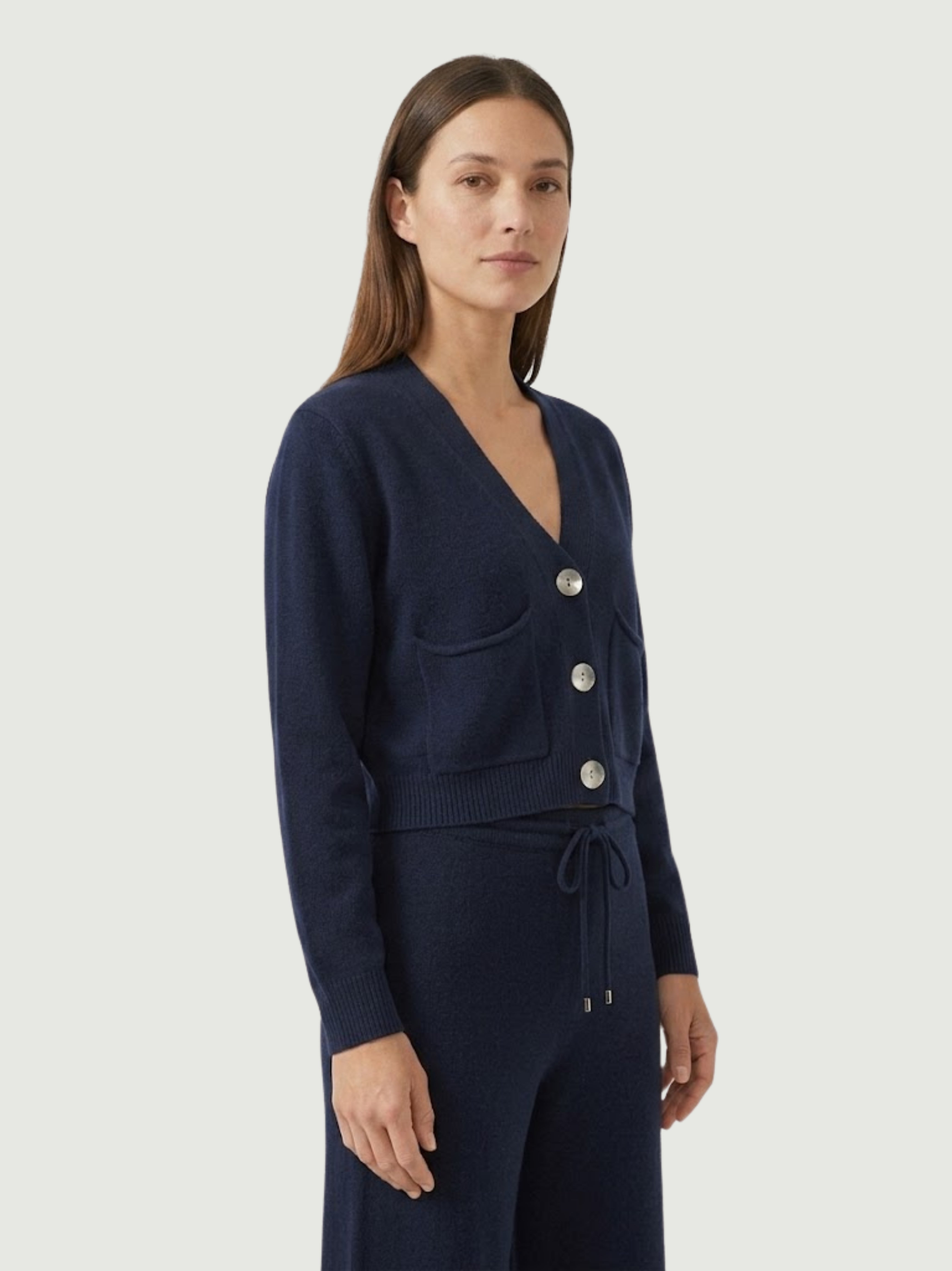 Vista laterale modella indossa cardigan cropped blu scuro con scollo a V, chiusura frontale con bottoni effetto madreperla, taschini applicati
