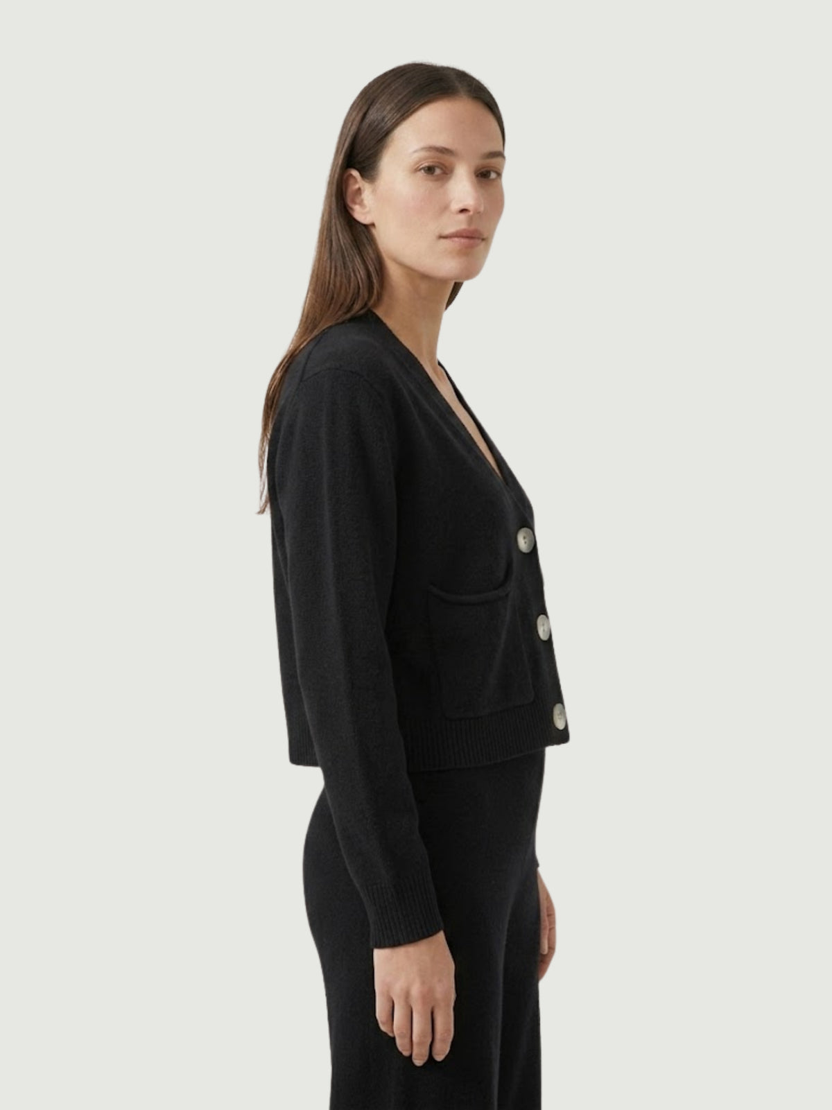 Vista laterale modella indossa cardigan cropped nero con scollo a V, chiusura frontale con bottoni effetto madreperla, taschini applicati