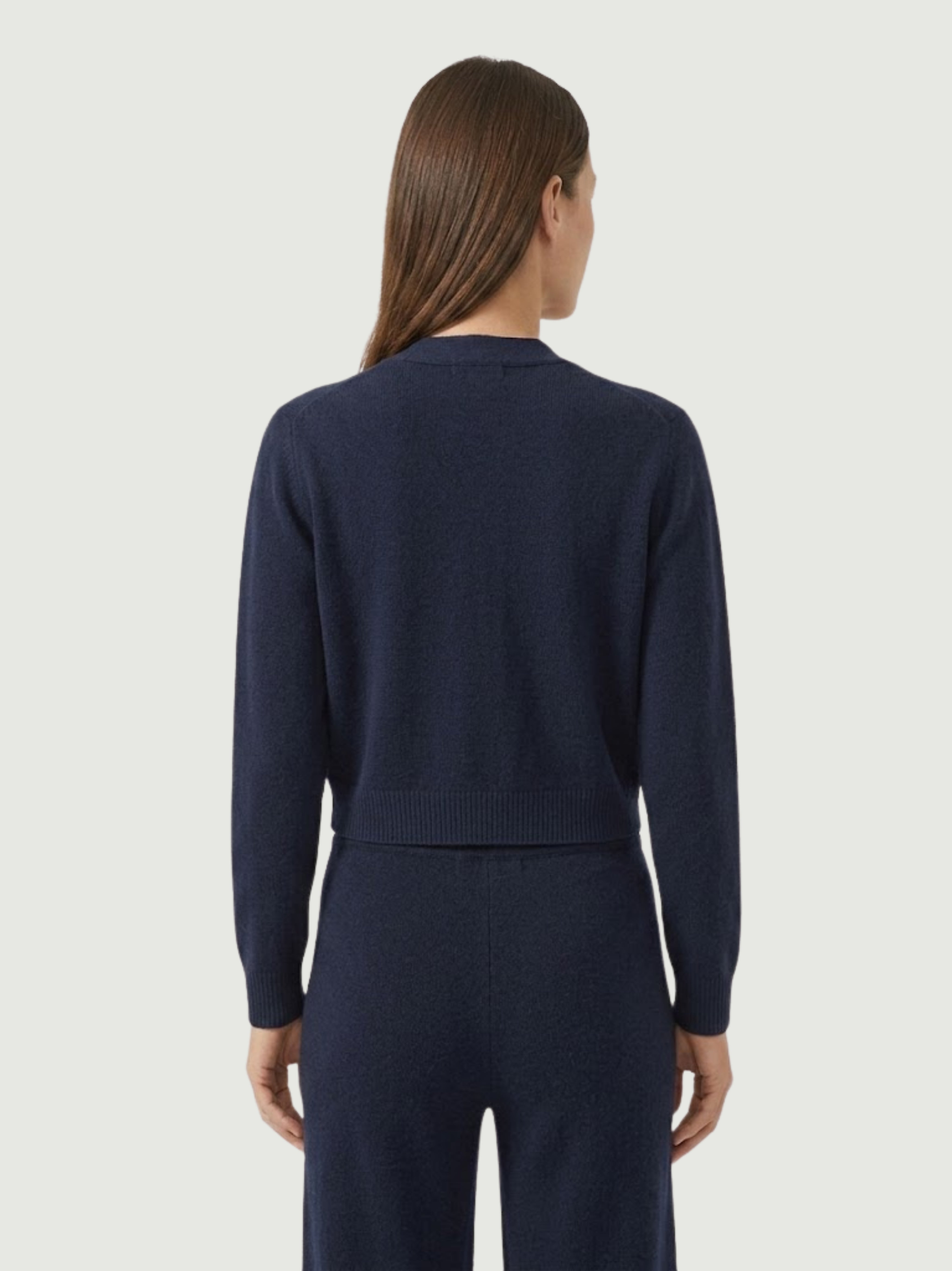 Vista posteriore modella indossa cardigan cropped blu scuro con scollo a V, chiusura frontale con bottoni effetto madreperla, taschini applicati